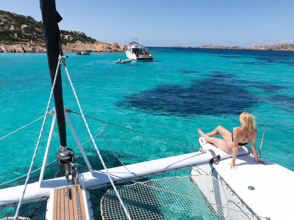 Woman in bikini tanning and relaxing on a summer sailin cruise, sitting on a luxury catamaran near picture perfect white sandy beach on Spargi island in Maddalena Archipelago, Sardinia, Italy.